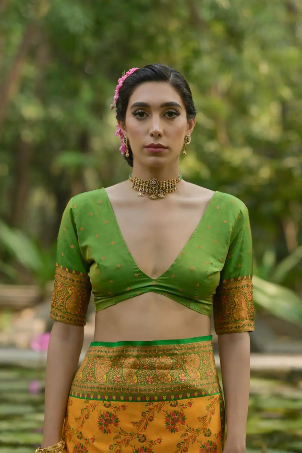 Woman wearing a mustard yellow saree with green blouse and intricate zari work, sitting by a stone water feature with blooming pink lotus flowers in a lush garden.
